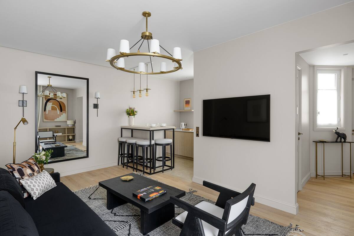 Una sala de estar moderna con un sofá negro, una mesa de centro, una araña y un televisor montado en la pared.