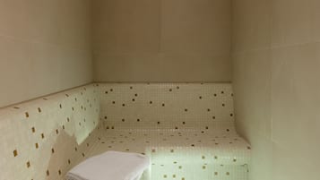 Un pequeño cuarto con baldosas, un banco, una toalla doblada y un par de pantuflas en el suelo.