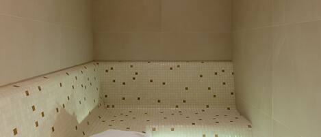 A small, tiled room with a bench, a folded towel, and a pair of slippers on the floor.