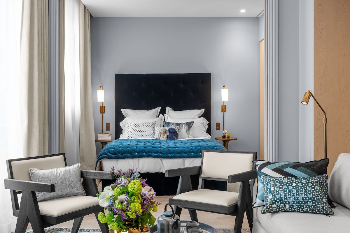 Una habitación de hotel moderna con una cama grande, dos mesitas de noche con lámparas, un sofá y una mesa de centro con flores.