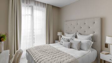 Habitación de hotel con una cama grande, una mesita de noche, una lámpara y vistas al exterior a través de cortinas translúcidas.
