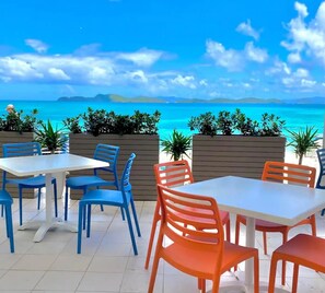 Un espace extérieur aménagé avec des tables et des chaises, offrant une vue sur une plage aux eaux d’un bleu limpide et sur la côte lointaine.