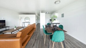 A modern living room with a brown leather sofa, a dining table with teal chairs, and a TV on the left.