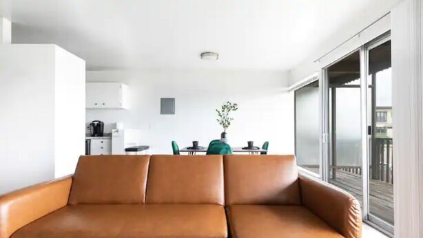 A modern living room with a brown leather sofa, a dining area with a green table and chairs, and a large sliding glass door leading to a balcony.