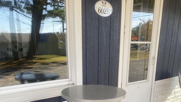 A gray table and two matching stools outside a building with a dark gray door and a window. A sign with the number "602B" is visible on the door. - Cute, cozy, updated cottage just steps from gorgeous Houghton Lake (Prudenville)