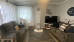 A living room with a grey sofa, a smaller grey armchair, a coffee table, a TV, and a ceiling fan. - Cute, cozy, updated cottage just steps from gorgeous Houghton Lake (Prudenville)