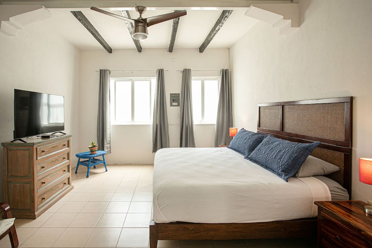 Une chambre avec un lit en bois, un téléviseur posé sur une commode et un ventilateur de plafond.