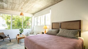 A bedroom with a large bed, a wooden headboard, a window with a view of greenery, a small table with a lamp, and a chair. - Casitas Miramar Puerto Vallarta Malecón (Puerto Vallarta)