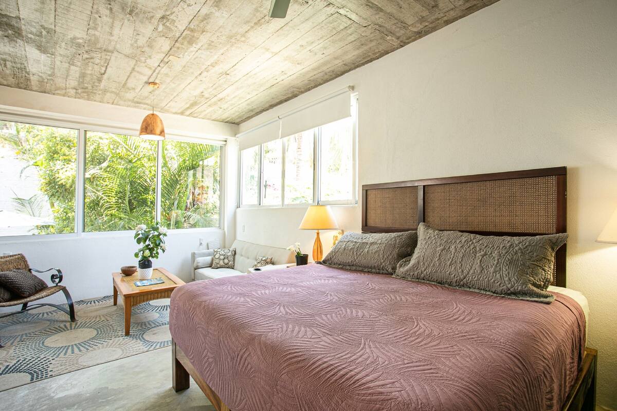 Une chambre avec un grand lit, une tête de lit en bois, une fenêtre donnant sur un paysage verdoyant, une petite table avec une lampe et une chaise.
