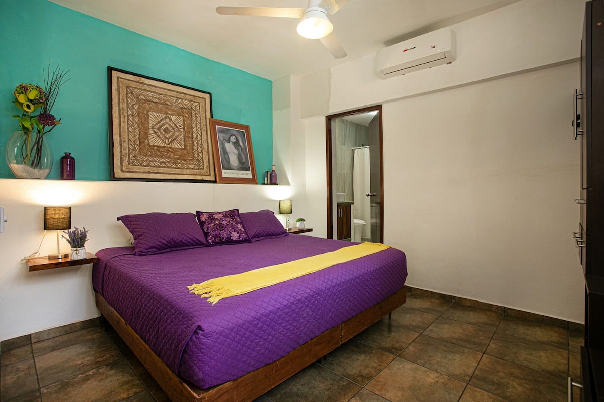 Une chambre à coucher avec un grand lit, un mur d’accent de couleur turquoise, un ventilateur de plafond et une porte donnant sur une salle de bain.