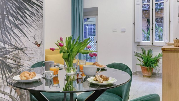 A dining area with a glass table, green chairs, breakfast items, a vase with flowers, and a view of the outside through a window.