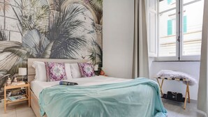 A bedroom with a large tropical mural, a bed with a teal bedspread, a nightstand with a lamp, and a window with a view of buildings.