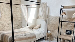 Un dormitorio con cama con dosel, una mesita de noche, una repisa con ropa y un dispositivo, y una alfombra estampada.