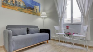 A modern room with a grey sofa, a small white table with chairs, a floor lamp, and a painting of a historical building on the wall. - APPARTAMENTO SANTI QUATTRO 2 - HOME AND MORE (Roma)