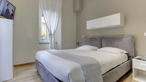 A neatly arranged bedroom with a bed, a bedside table, a TV mounted on the wall, and a window with curtains. - APPARTAMENTO SANTI QUATTRO 2 - HOME AND MORE (Roma)