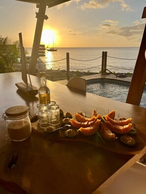 A table with a sunset view, a bottle, glasses, and a fruit platter.