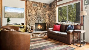 A cozy living room with a stone fireplace, leather furniture, and a large window with a view of trees. - Monterey Log Lair With Pool Table - Slope Views (Big Bear Lake)