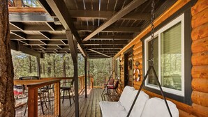 A wooden deck with a swing chair and a cushioned bench, overlooking a forested area. - Monterey Log Lair With Pool Table - Slope Views (Big Bear Lake)
