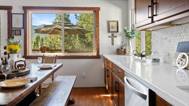 A kitchen with wooden cabinets, a large window with a view of trees, a dining table with chairs, and a white countertop. - Monterey Log Lair With Pool Table - Slope Views (Big Bear Lake)