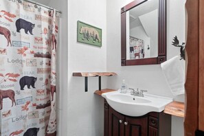 A bathroom with a wooden vanity, a white sink, a mirror, and a shower curtain with animal prints. - Monterey Log Lair With Pool Table - Slope Views (Big Bear Lake)