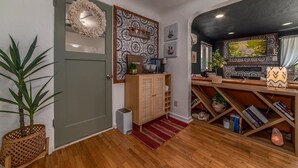 Um corredor com piso de madeira, porta verde, placa de boas-vindas, parede estampada e uma estante com livros e objetos decorativos.