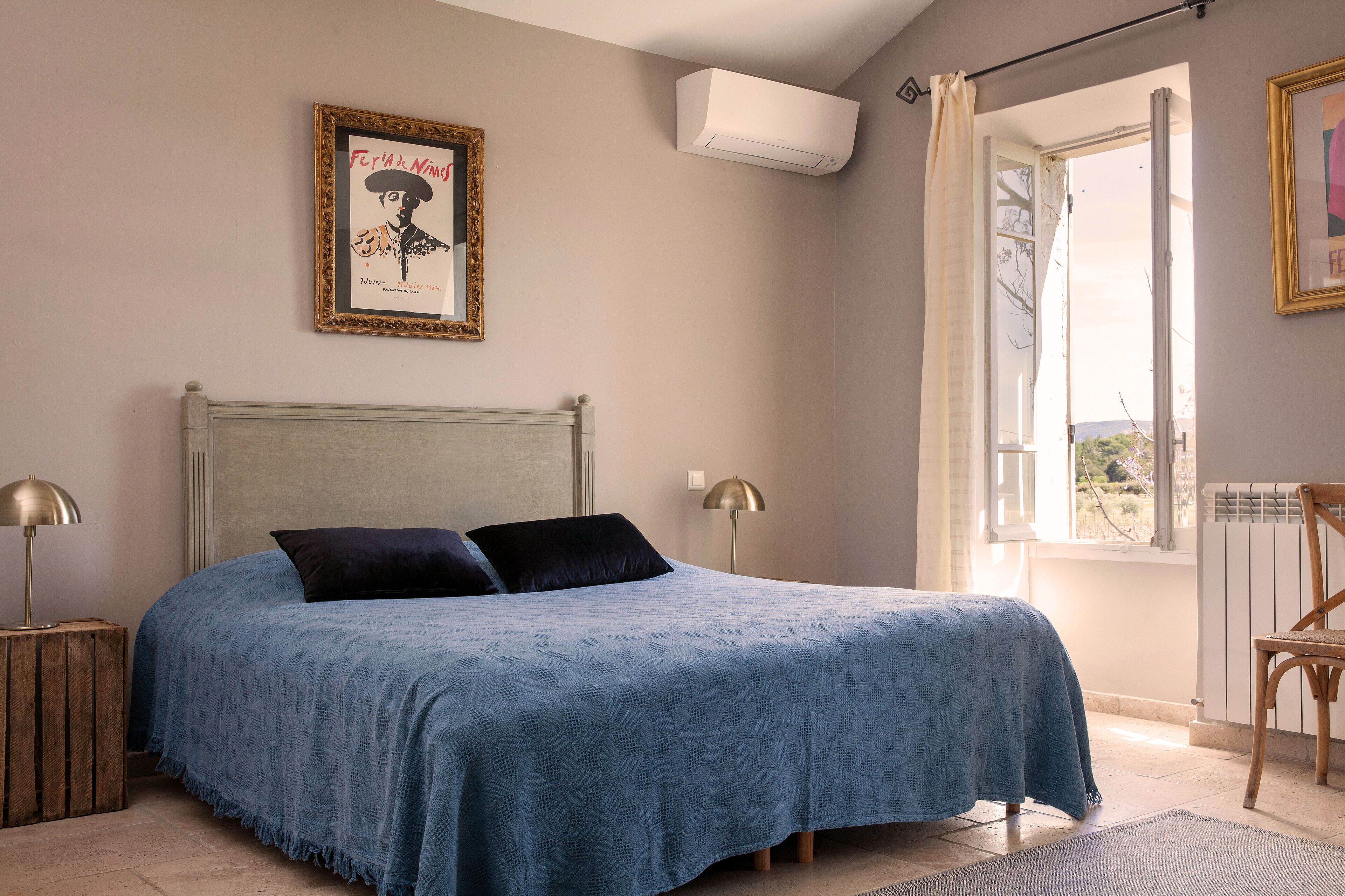 Une chambre à coucher avec un lit, des lampes de chevet, un tableau encadré, un climatiseur et une fenêtre avec des rideaux.
