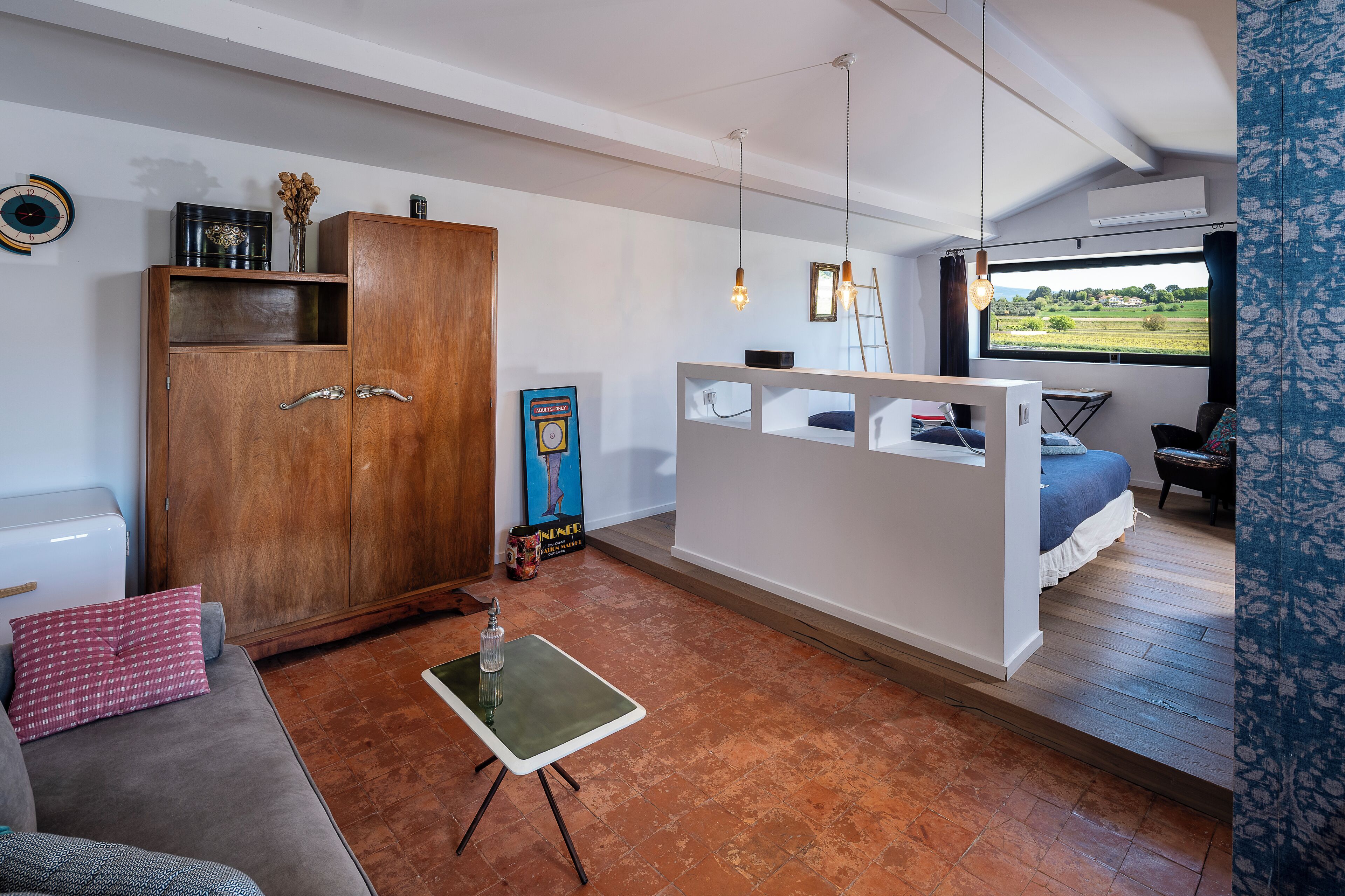 A compact room with a wooden wardrobe, a small table, a sofa, a bed with a view, and a window.