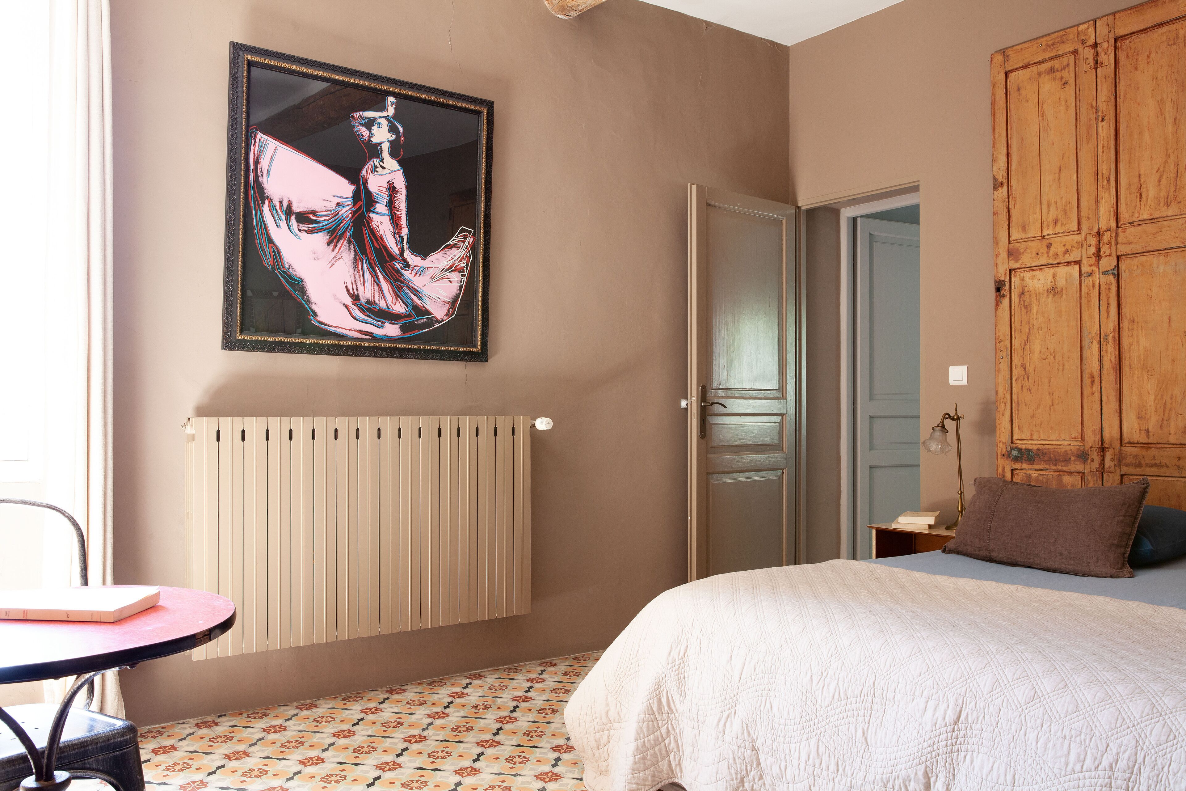 Ein Schlafzimmer mit einem Bett, einem Heizkörper, einem Holzschrank, einem Tisch mit Stuhl und einem gerahmten Kunstwerk an der Wand.