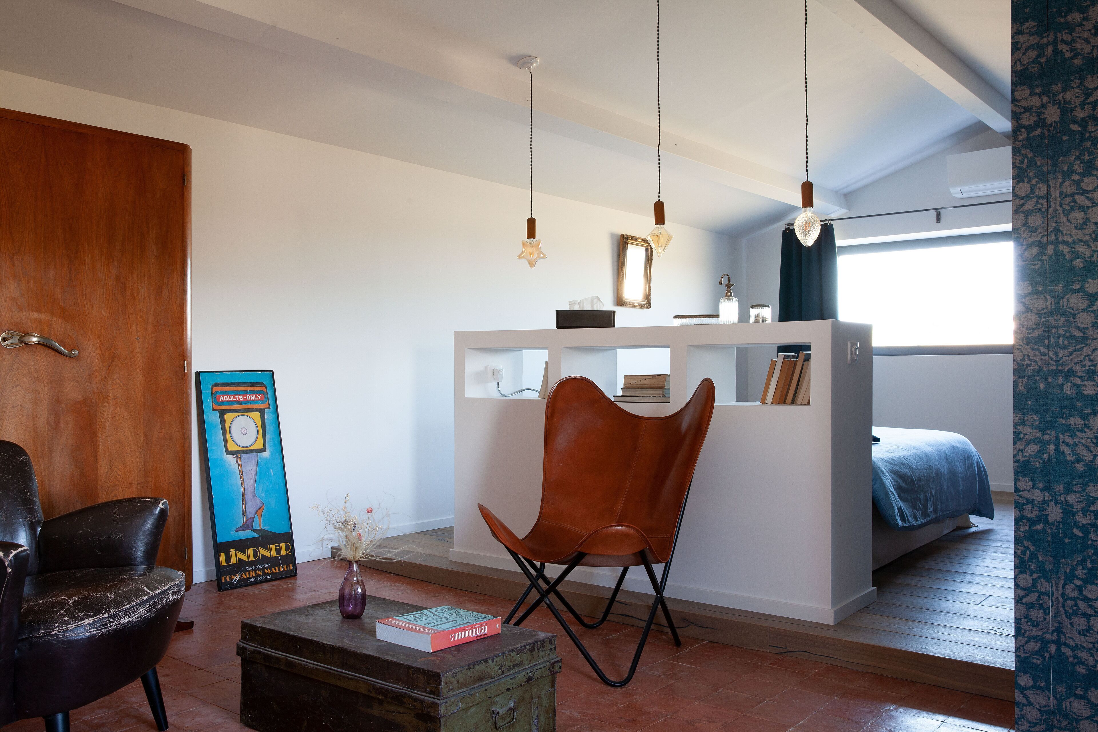 Una habitación moderna con suelo de madera, una cama blanca, un sillón de cuero marrón, una mesita de consola blanca y un cartel grande en la pared.