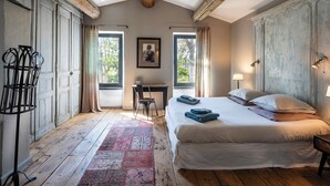 A bedroom with a large bed, a desk, and a chair. There are windows with curtains, a wooden floor, and a ceiling with exposed beams.