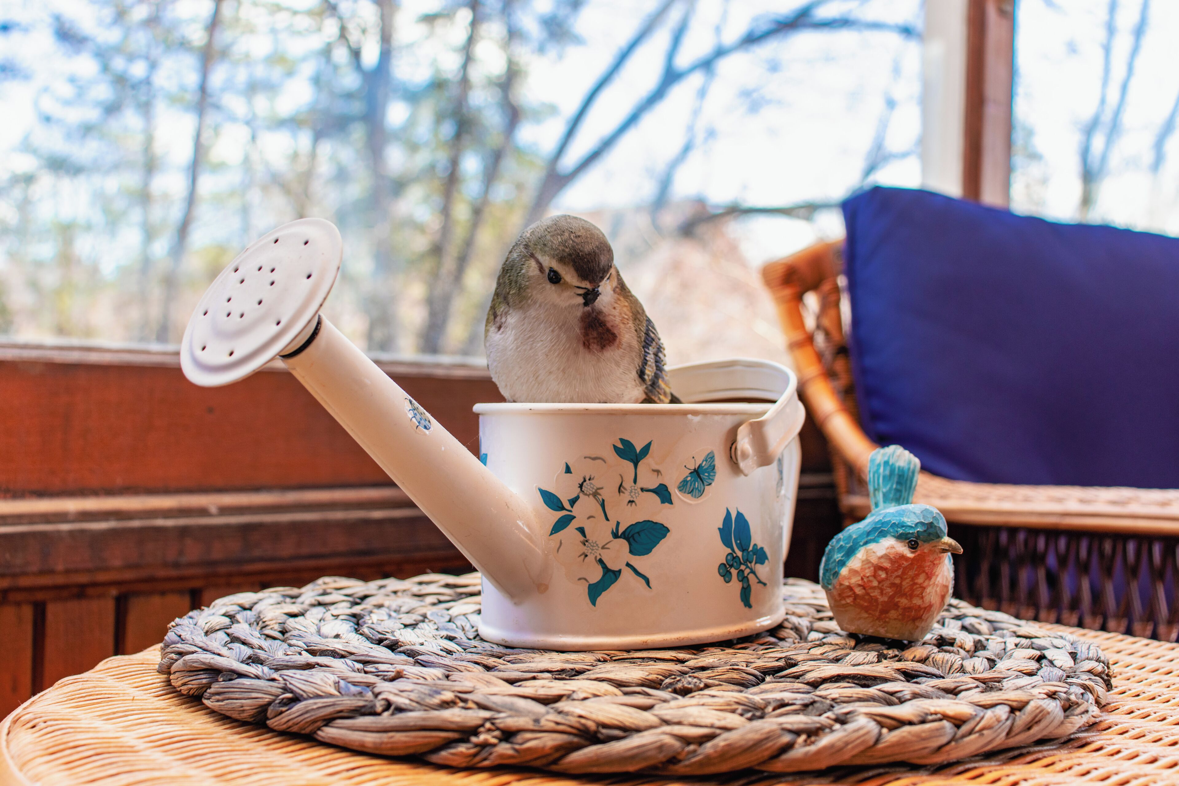 Un pájaro posado sobre una tetera con diseños florales, sobre un tapete tejido.