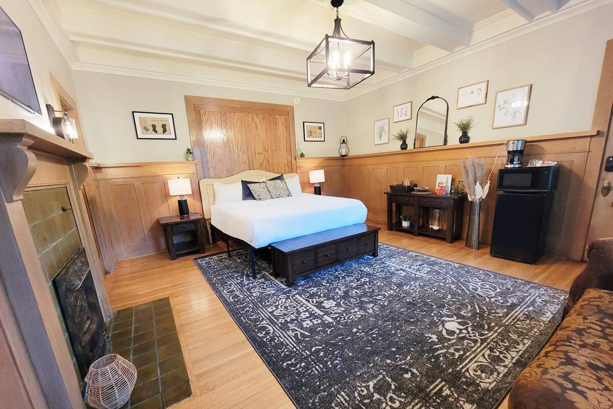 A bedroom with a bed, bedside tables, a nightstand, a dresser, a mirror, a fireplace, and a rug.