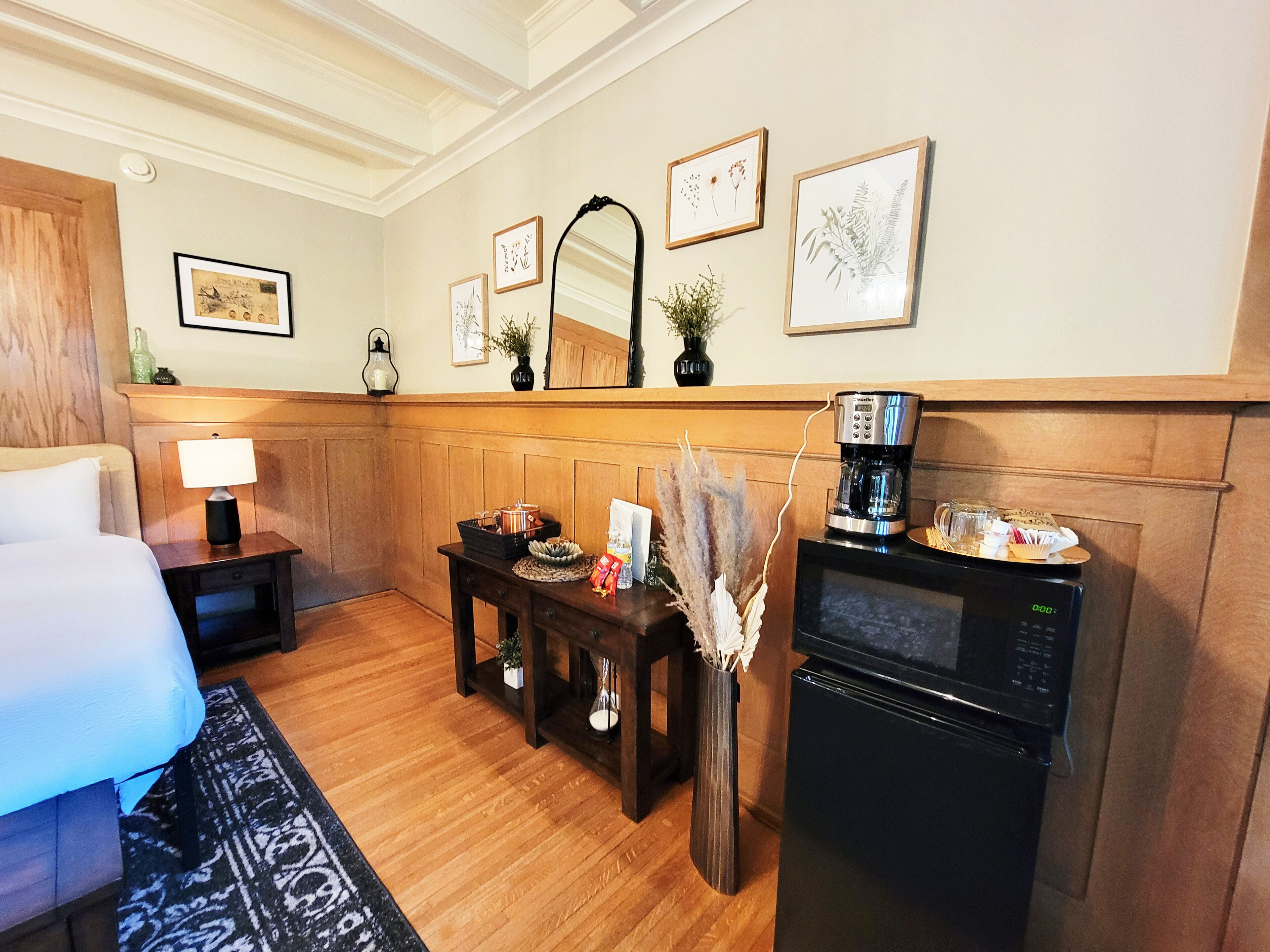 Una habitación de hotel con una cama, una mesita de noche con una lámpara, una pequeña mesa con un jarrón de flores secas, una cafetera y un refrigerador.