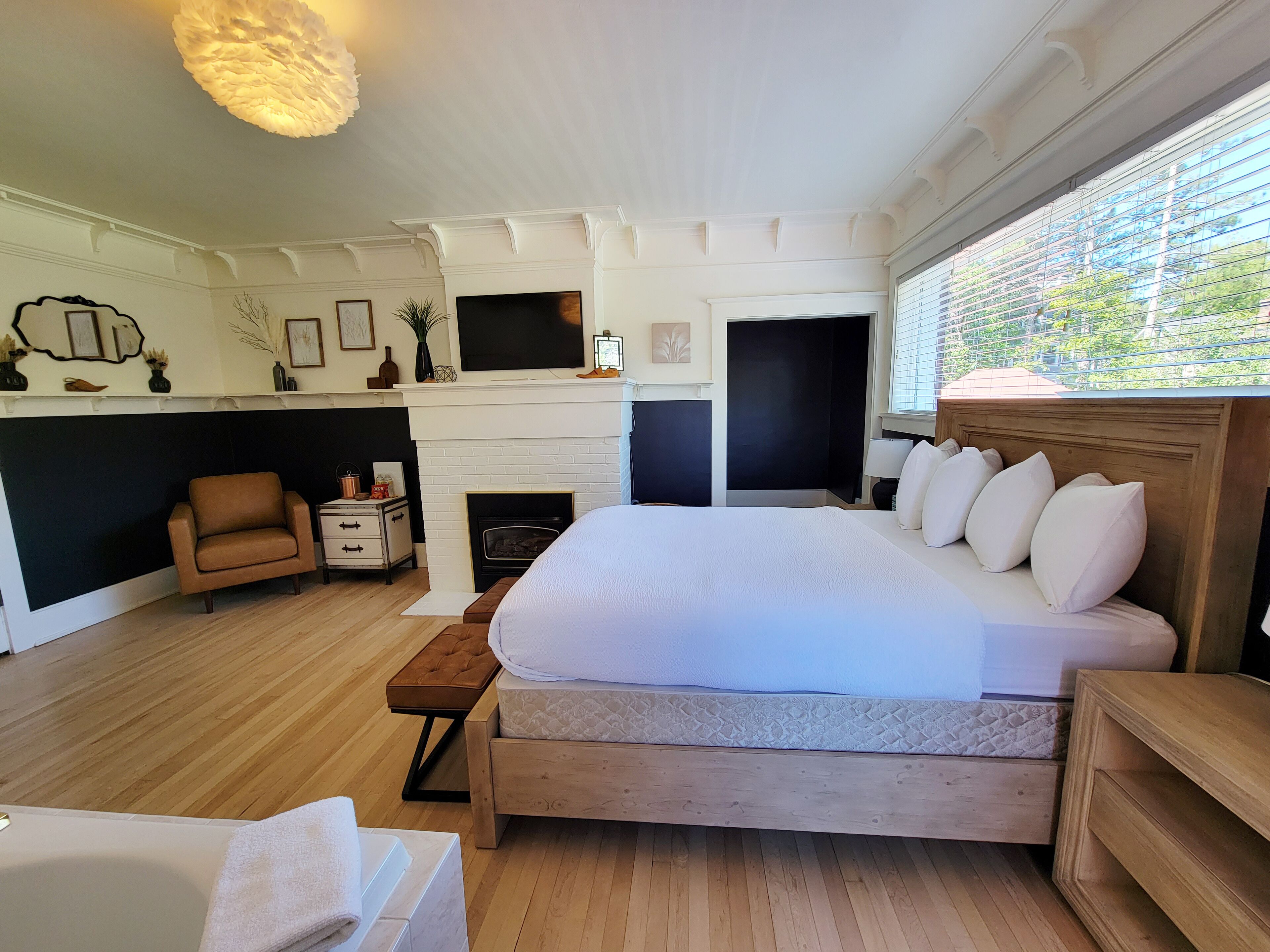 A bedroom with a large bed, a wooden nightstand, a small table, a chair, a fireplace, and a TV.