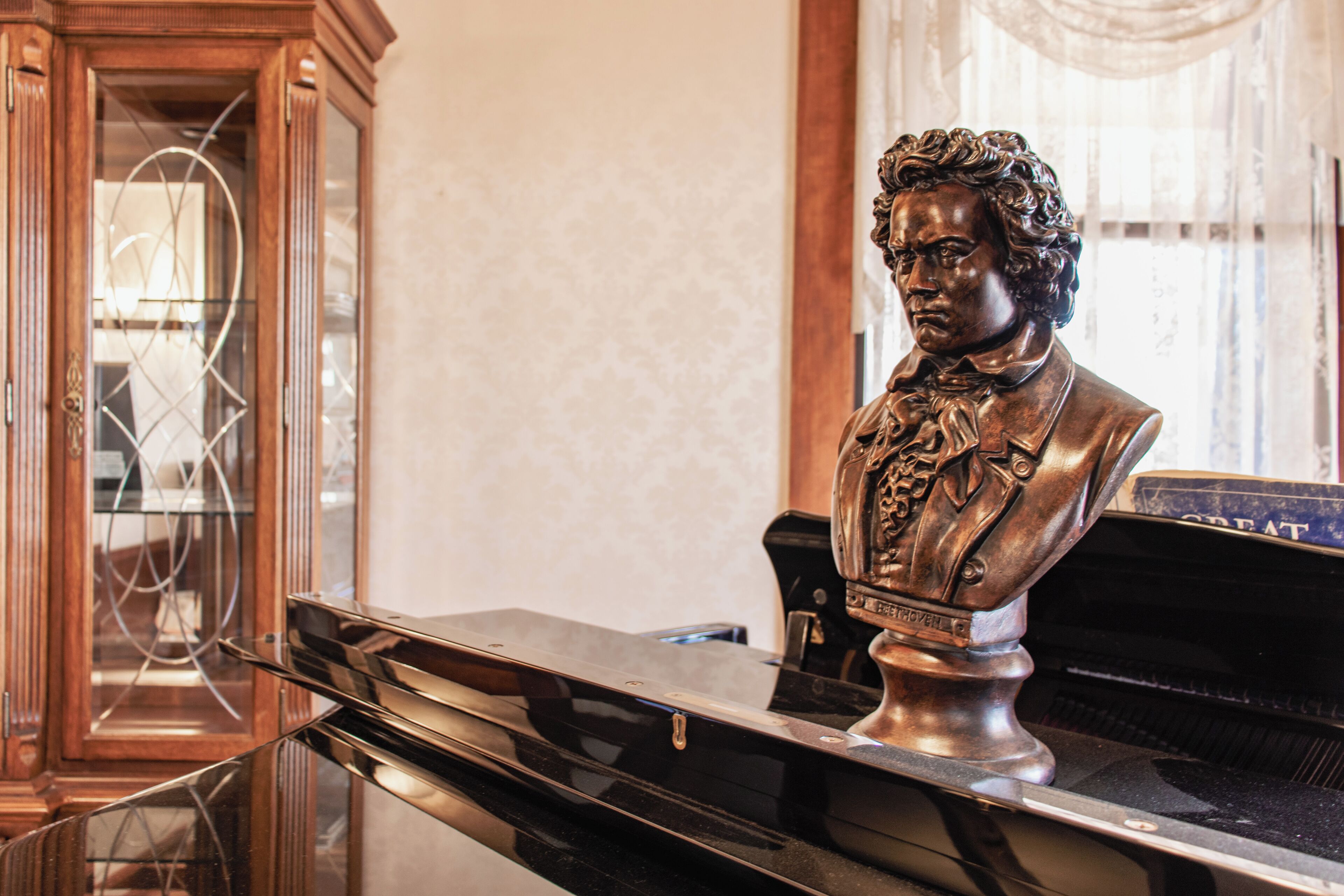 A bust of a person on a piano in a room with a wooden cabinet and a window with curtains.