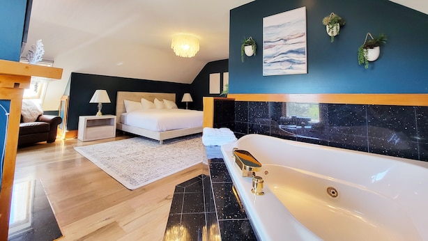 A modern bedroom with a large bed, a sitting area with a chair, a small table, and a bathtub. - Historic GG Barnum Mansion by Heirloom Boutique Collection (Duluth)