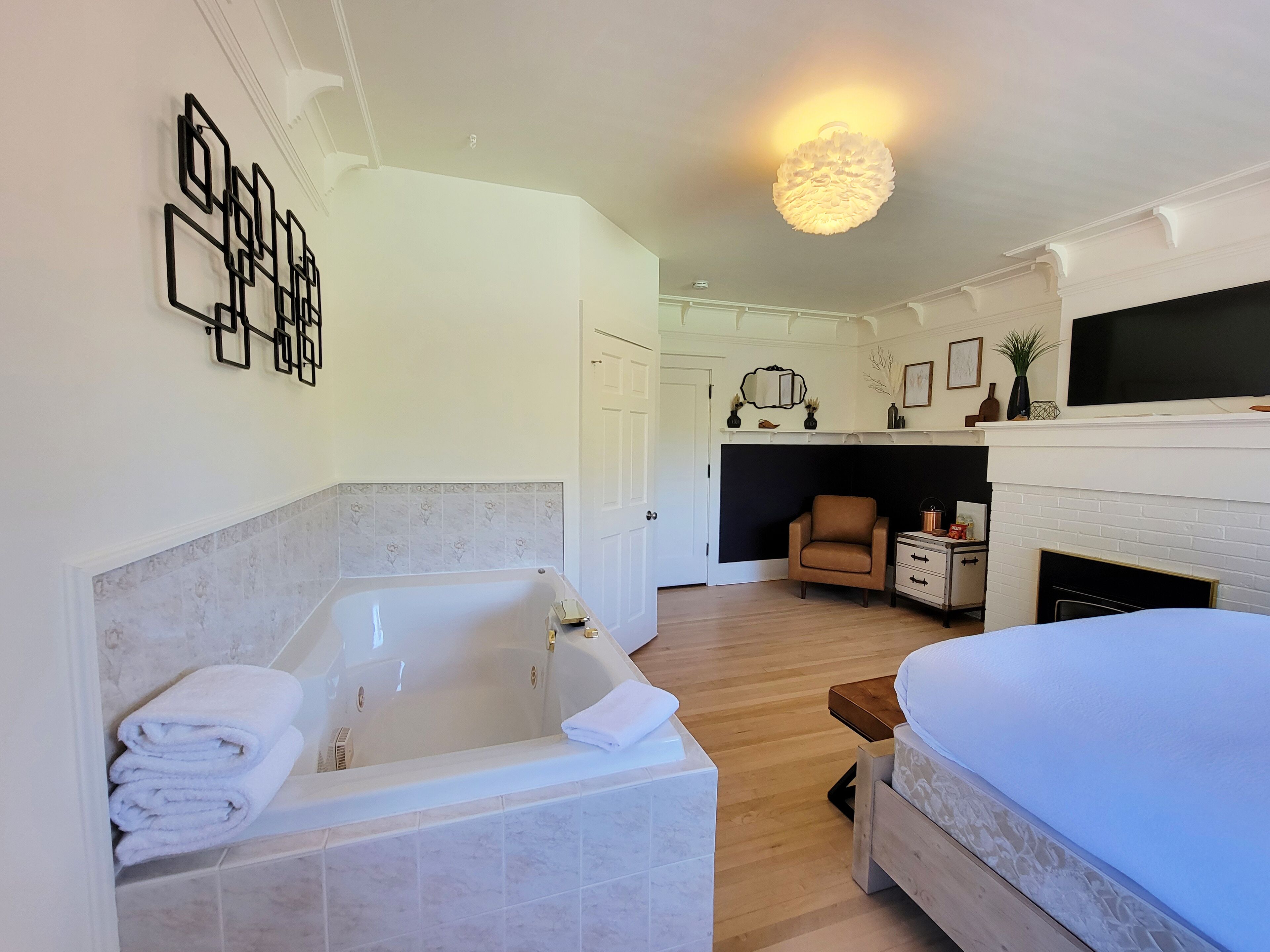 A modern hotel room with a large bathtub, a bed, a fireplace, and a television.