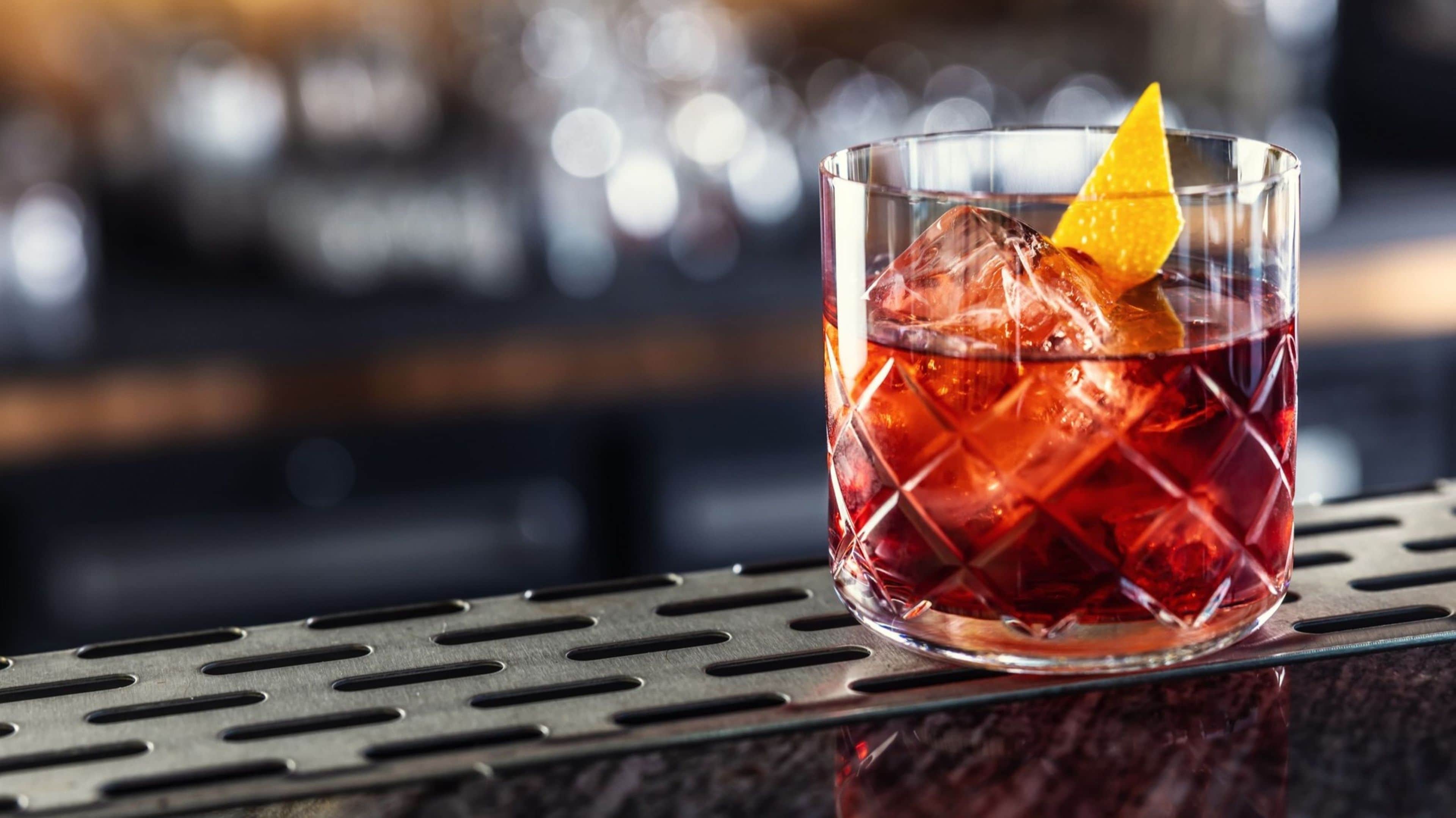 A glass with a red beverage, ice cubes, and a lemon slice on a metal bar counter.