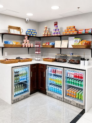 A self-service beverage station with refrigerated drink dispensers and shelves stocked with snacks and beverages. - MONTREAL SUITES ORLANDO by IHG (Orlando)