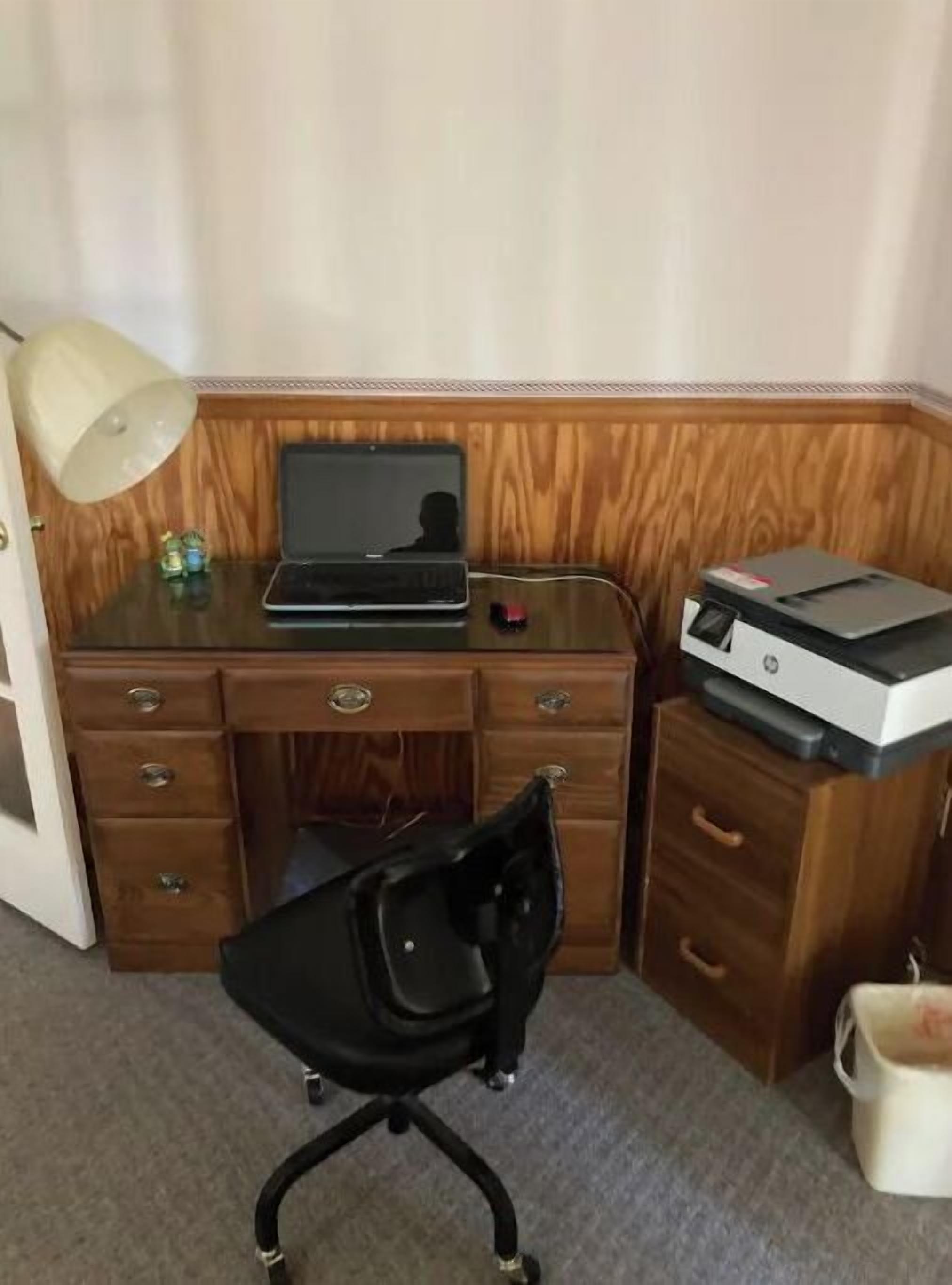 Ein Büro mit Eckausrichtung, ausgestattet mit einem Holztisch, einem Laptop, einem Drucker, einem schwarzen Bürostuhl, einer Lampe und einem kleinen dekorativen Gegenstand.