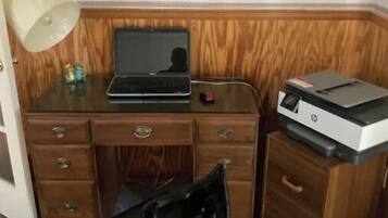 Ein Büro mit Eckausrichtung, ausgestattet mit einem Holztisch, einem Laptop, einem Drucker, einem schwarzen Bürostuhl, einer Lampe und einem kleinen dekorativen Gegenstand.