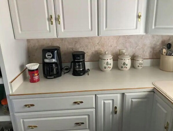 Una cocina con armarios blancos, una cafetera, un recipiente rojo y dos tarros blancos con tapa.