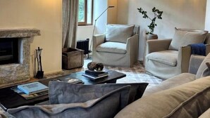 Ein Wohnzimmer mit Kamin, zwei Sesseln, einer Couch und einem Couchtisch. An den Wänden sind Leuchten angebracht und es gibt ein Fenster mit Vorhängen.