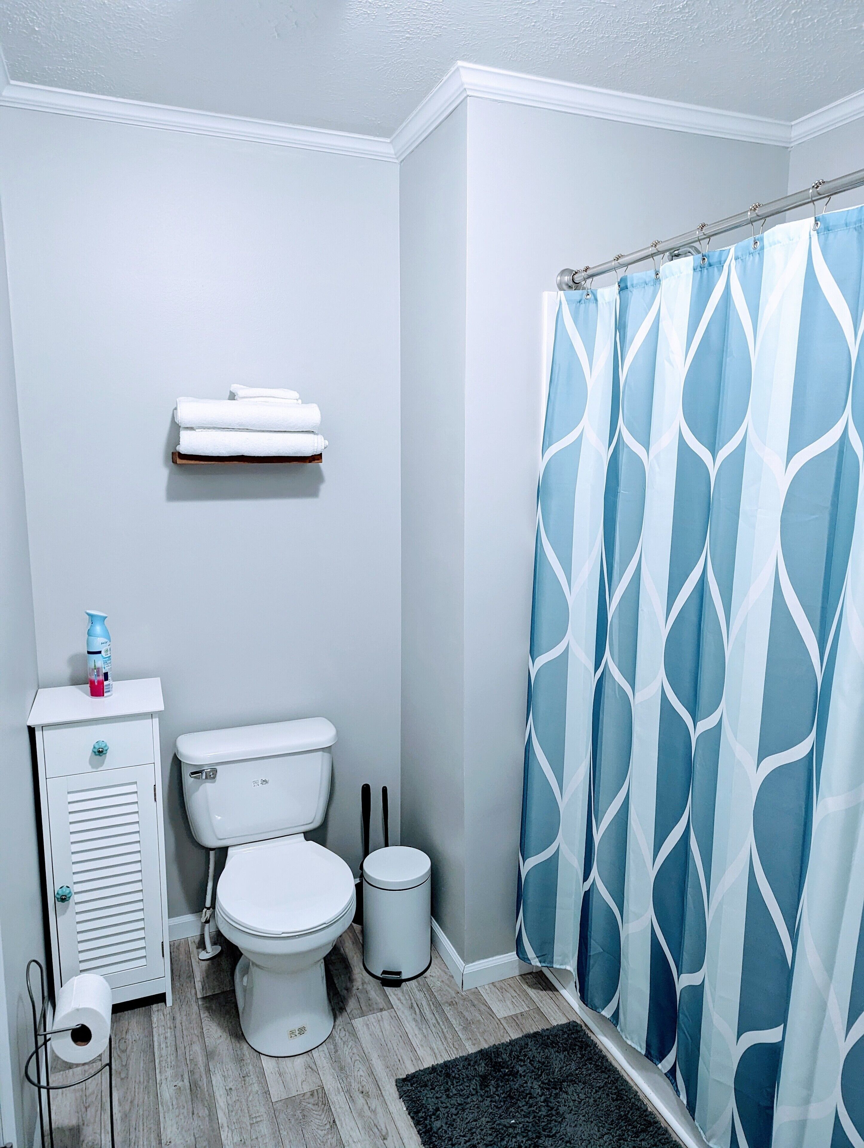 Une salle de bain comprenant des toilettes, un meuble blanc, une poubelle, un rideau de douche à motifs bleus et blancs, et une petite étagère au-dessus des toilettes.