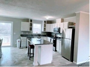 A modern kitchen with white cabinets, a central island, stainless steel appliances, and a large window providing natural light.