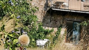 A rustic stone building with a balcony, surrounded by overgrown vegetation and a table set outdoors. - Centaurée** - Charm and countryside at a Witch on the Roof (Firmi)
