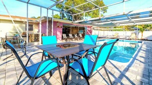 Een buitenzwembad met een overdekt terras, blauwe stoelen en een tafel.