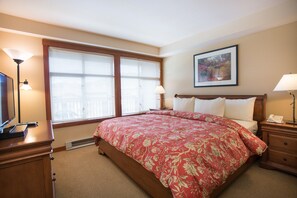 A bedroom with a large bed, two bedside tables, a nightstand, a window with blinds, a wall-mounted painting, and a television on a dresser.