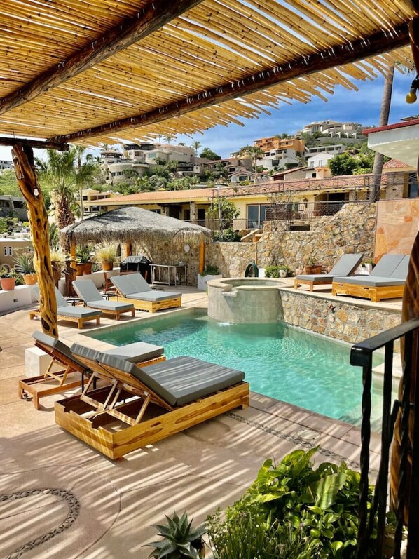 A pool area with lounge chairs, a hot tub, and a thatched roof structure.