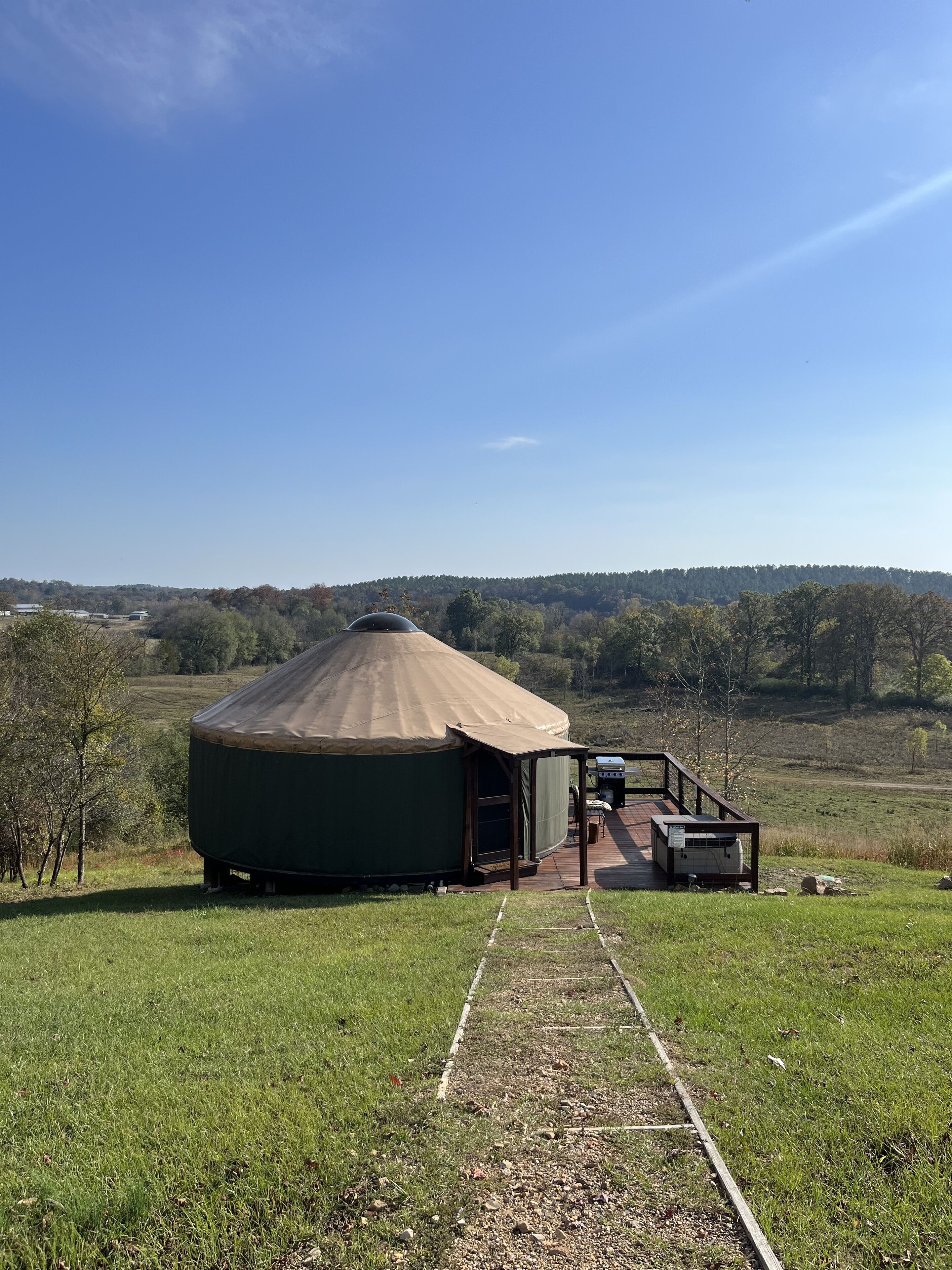 A yurt with a covered porch and a small deck, surrounded by a grassy field and trees.